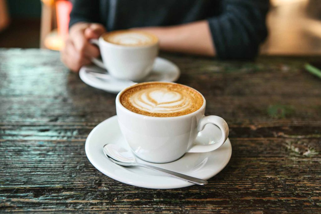Two cups of coffee with latte art on a wooden table, one cup in the foreground and a hand holding another in the background.