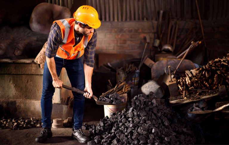 A man in a hard hat and safety vest shovels coal in a workshop filled with tools and materials.