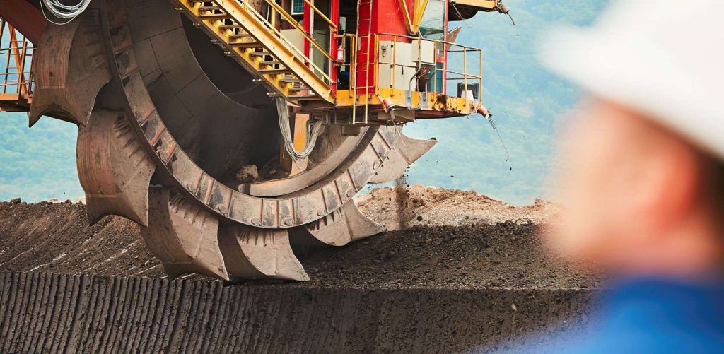 A large mining machine with a circular blade is excavating earth, with a worker in a hard hat blurred in the foreground.