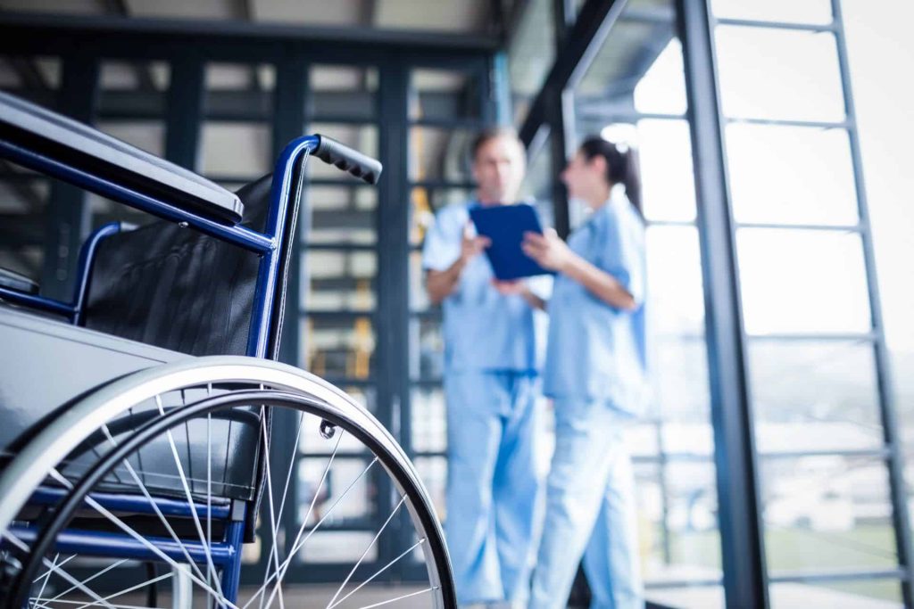 A wheelchair is in the foreground with two healthcare professionals discussing in the background.