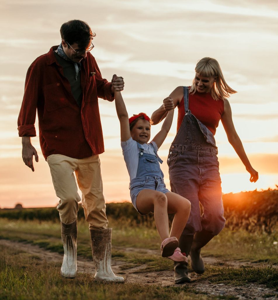 Parents swinging their young daughter between them on a country track at sunset.