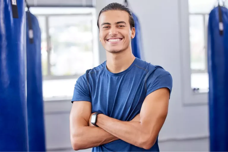 Smiling young man in a blue shirt stands confidently with arms crossed in a gym setting.