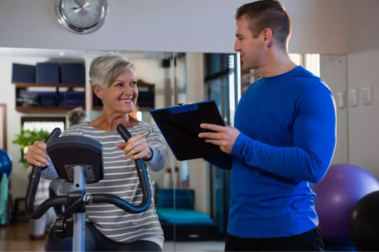 A smiling older woman exercises on a stationary bike while a young man holds a clipboard and talks to her.