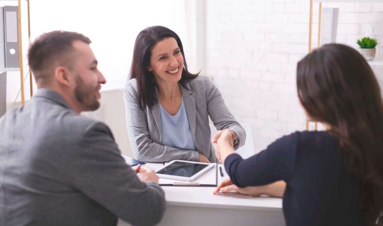 A woman in a blazer smiles while shaking hands with a client at a desk, with a man sitting beside her.