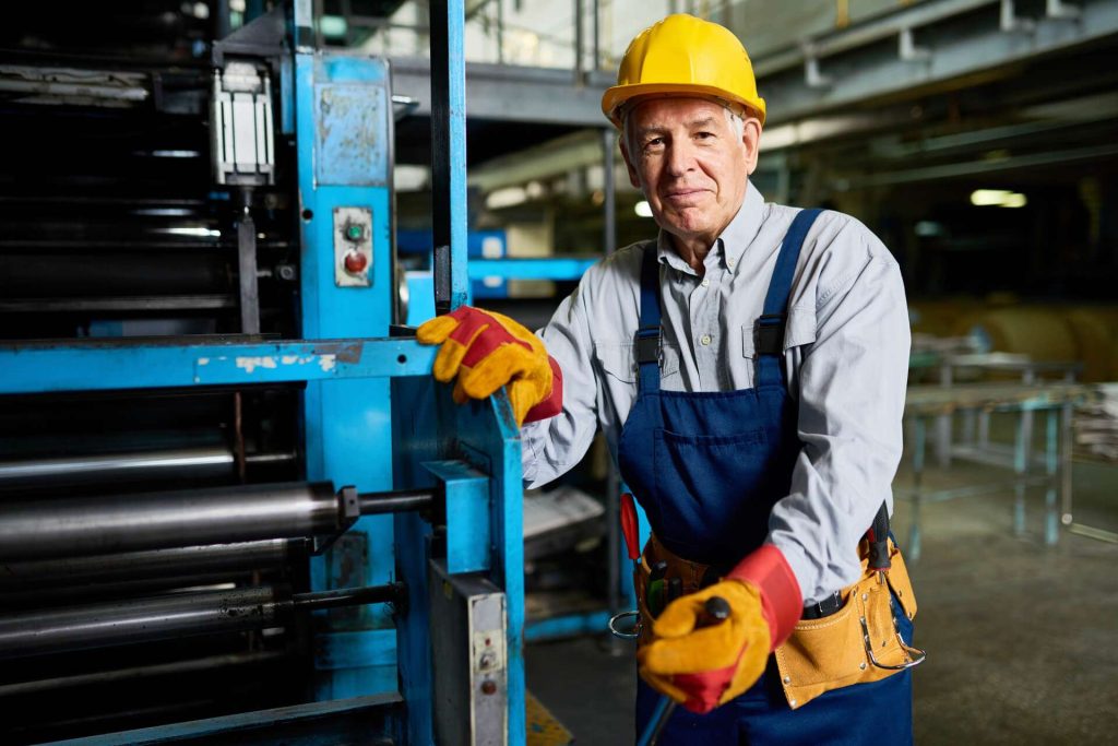 A worker in a hard hat and gloves stands beside industrial machinery in a factory setting.