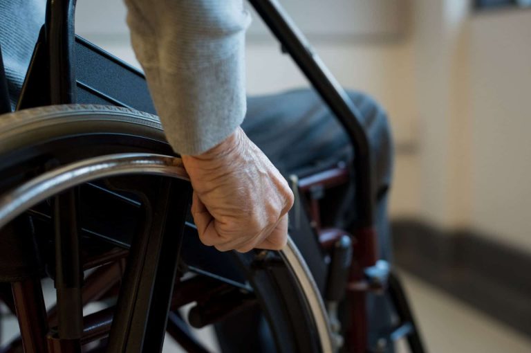A hand grips the wheel of a wheelchair, with a blurred background suggesting an indoor setting.