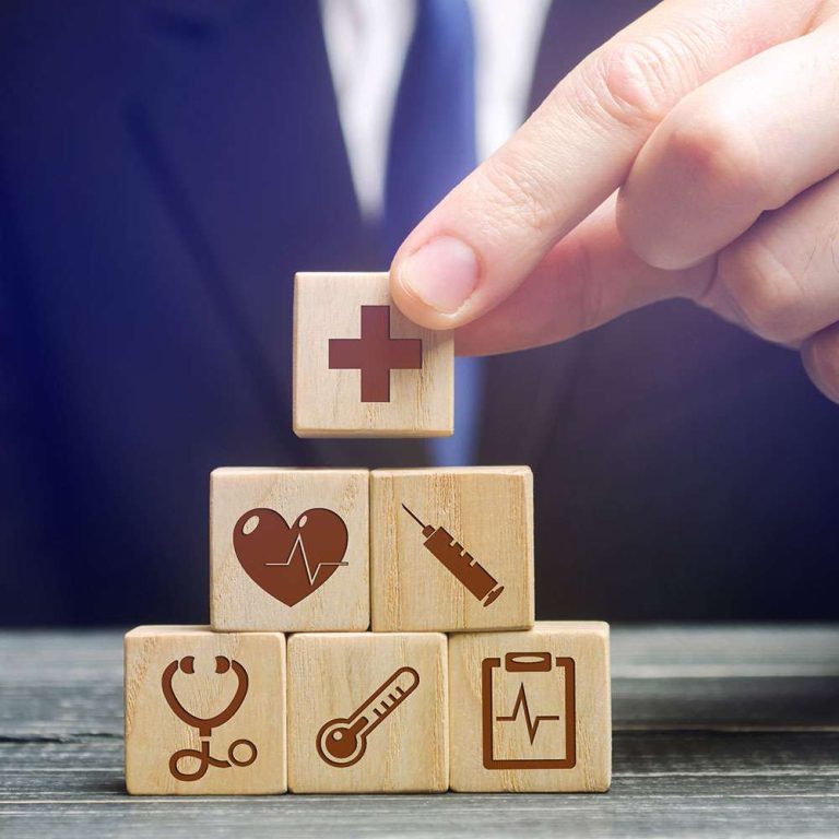 A hand places a wooden block with a medical cross on top of stacked blocks featuring health-related icons.