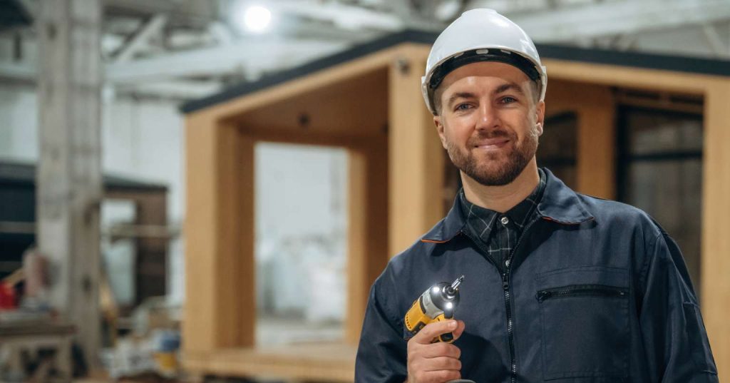 A smiling worker in a hard hat holds a drill in a construction setting with a partially built structure in the background.
