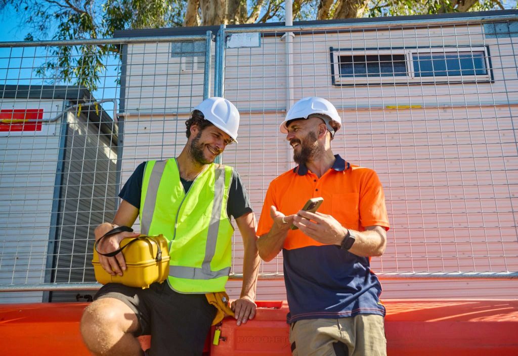 Two construction workers in safety gear are smiling and chatting while sitting on a barrier at a worksite.