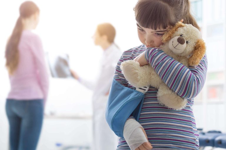 A young girl with a broken arm hugs a stuffed dog while looking concerned in a medical setting.