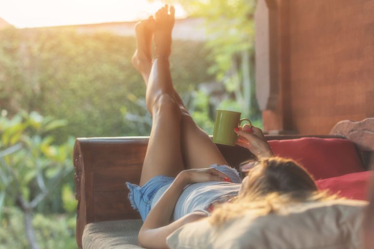 A person relaxes on a couch with their feet up, holding a green mug in a sunlit outdoor setting.